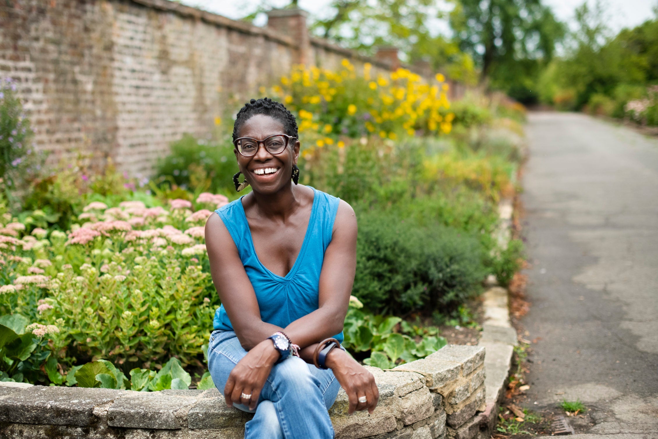 Flo Headlam sitting on a wall of a garden filled with colourful shrubs and plants