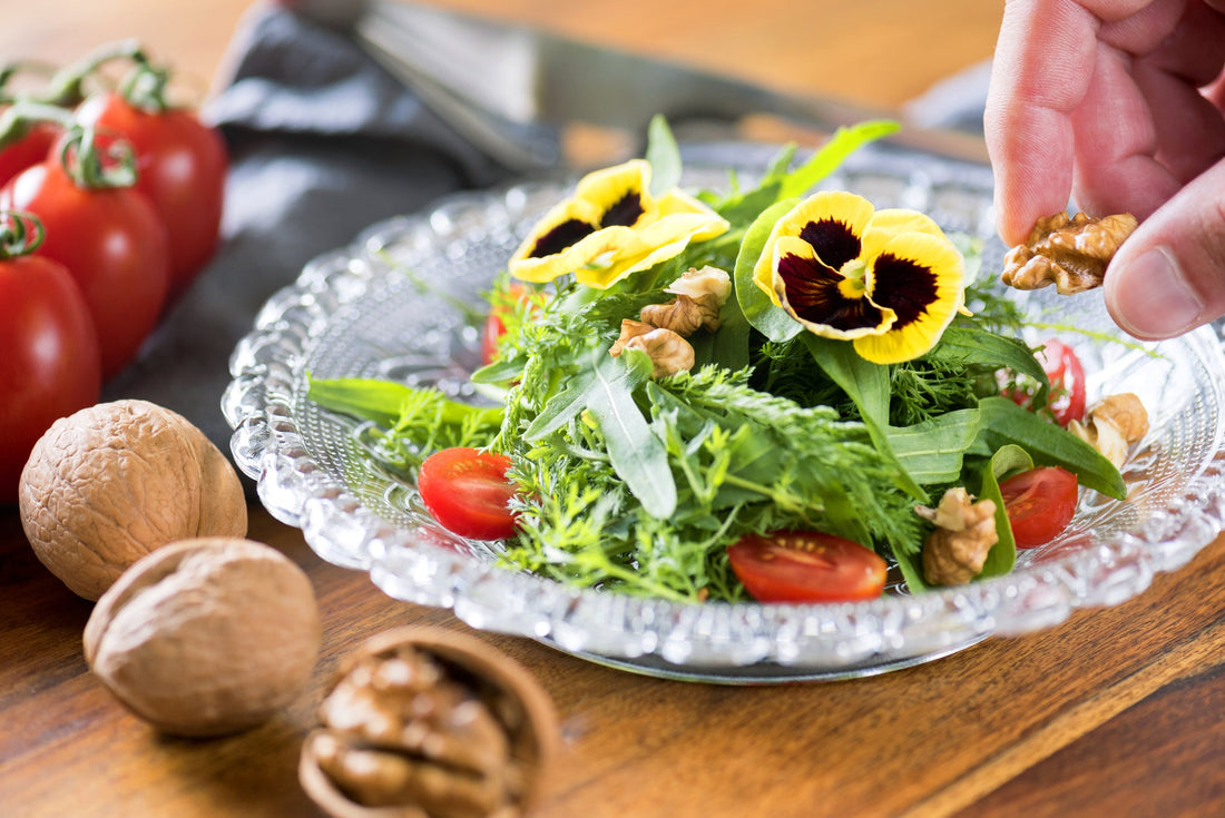 petals from a yellow and purple pansy flower used as a garnish on top of a green leafy tomato salad with nuts on the table next to it