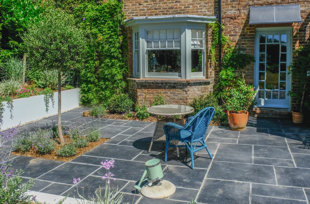 blue slate patio at the back of a house with a tree, plants in pots and small garden furniture