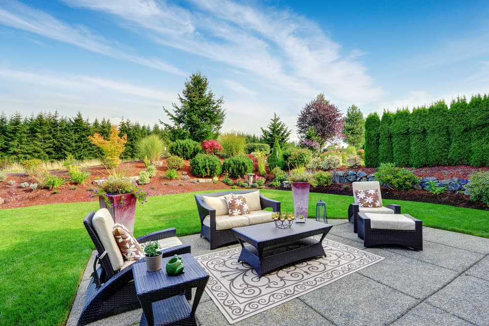 a cosy patio area with outdoor rug on stoned pavers, with a surrounding lawn and various trees and shrubs