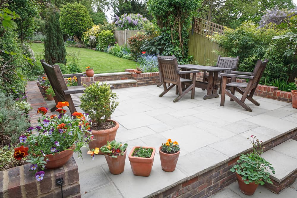a fenced off patio area with steps leading up a slope to a lawned area and plants including pansies and marigolds growing in pots
