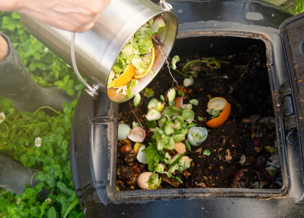 a home composting bin full of vegetables and other food waste being emptied into a larger outdoors bin, with a person wearing wellies in the background