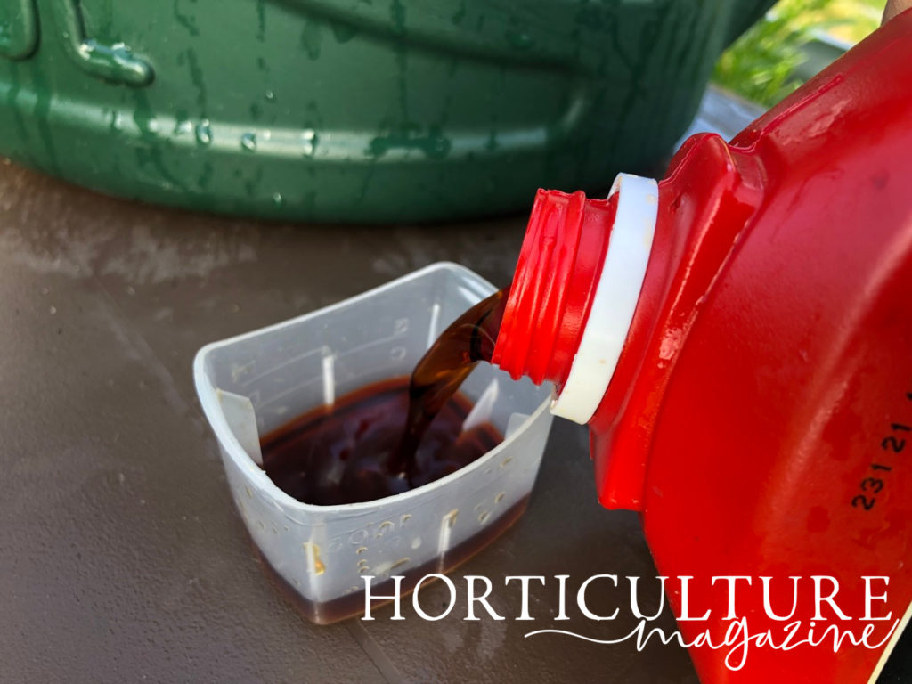 liquid feed being poured from a red bottle into a clear tub on a table with a watering can in the background