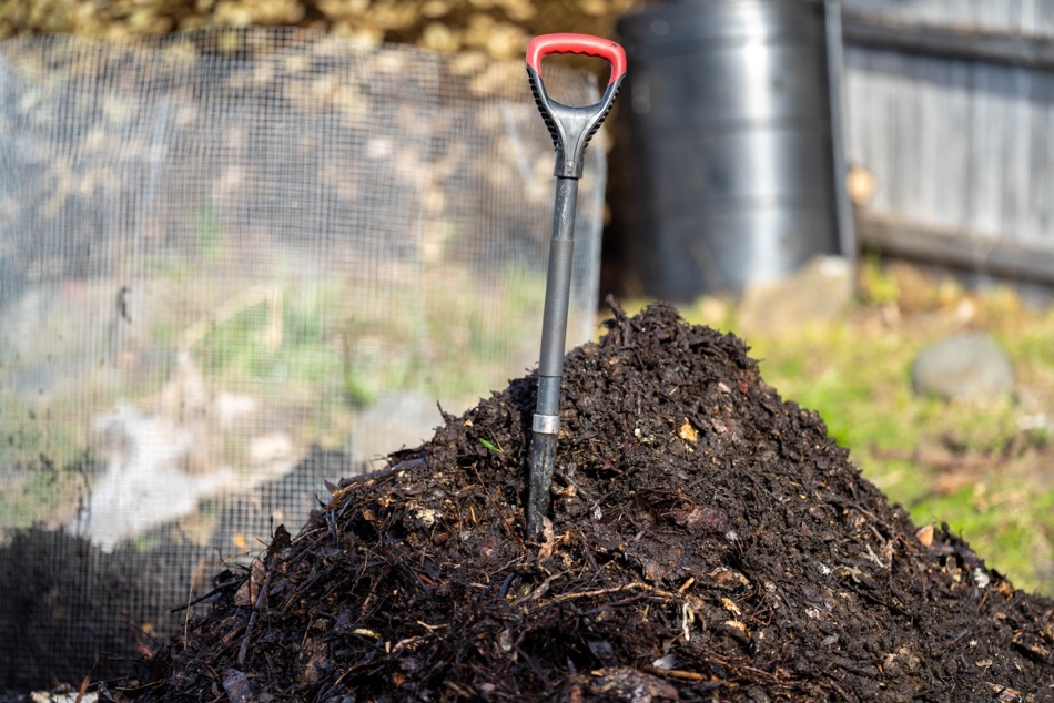 a large compost pile with a shovel stuck into it with a garden and netting in the background