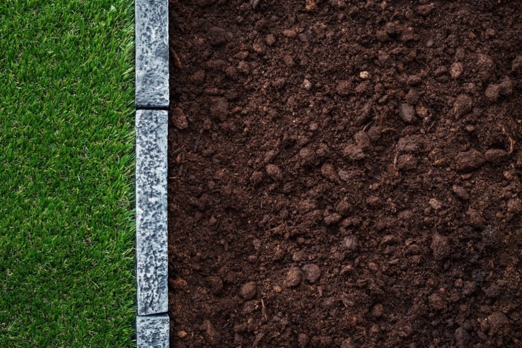 a garden bed full of compost next to a strip of green grass, divided by some garden slabs