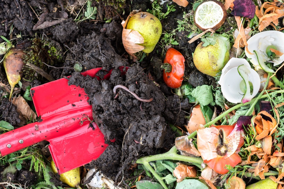 a red spade digging into some fresh compost that is full of food waste, such as carrots, eggshells, onions, peppers, amongst other things