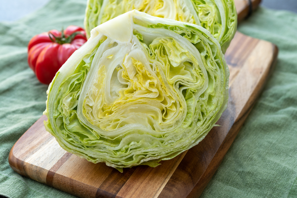 harvested iceberg lettuce cut in half on a chopping board with a tomato behind it