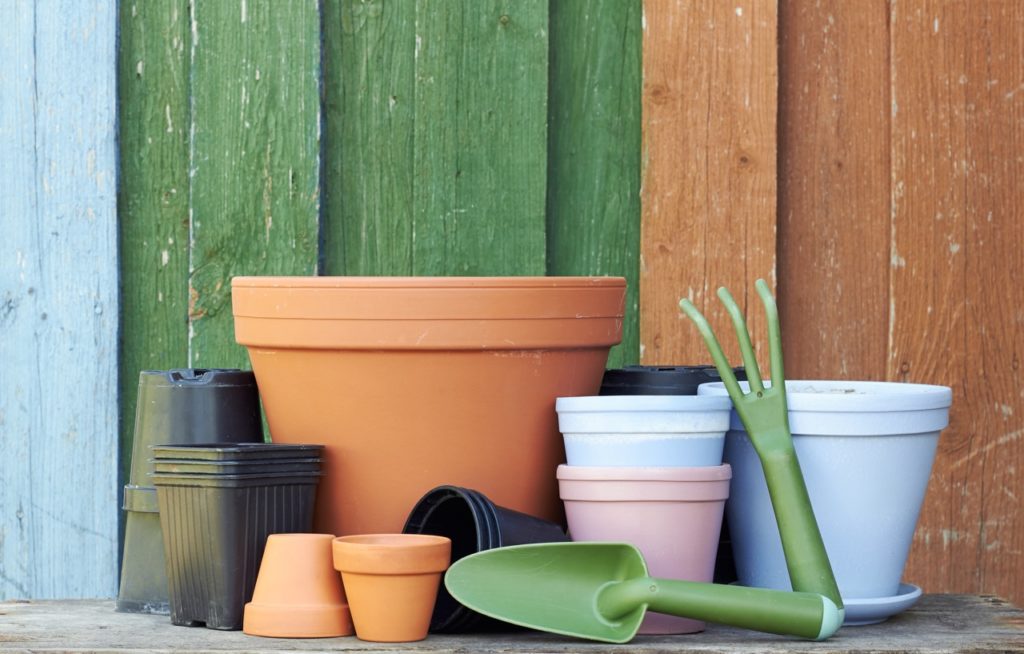 different containers of all shapes and sizes and colours, with a couple of garden tools in front of them placed outside in front of a green, brown and blue garden fence