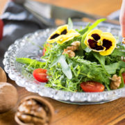 petals from a yellow and purple pansy flower used as a garnish on top of a green leafy tomato salad with nuts on the table next to it