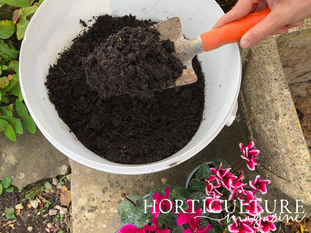 a trowel placing soil into a large container that is already half-full with compost