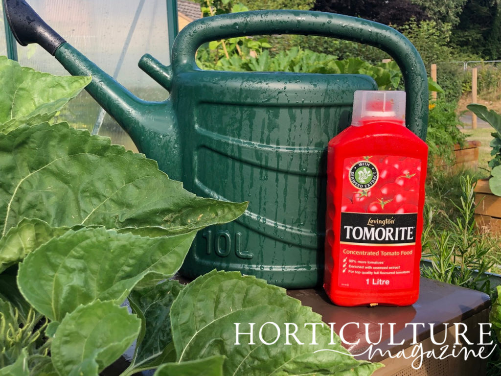 a watering can next to a bottle full of fertiliser on a table outside next to large green leaves 