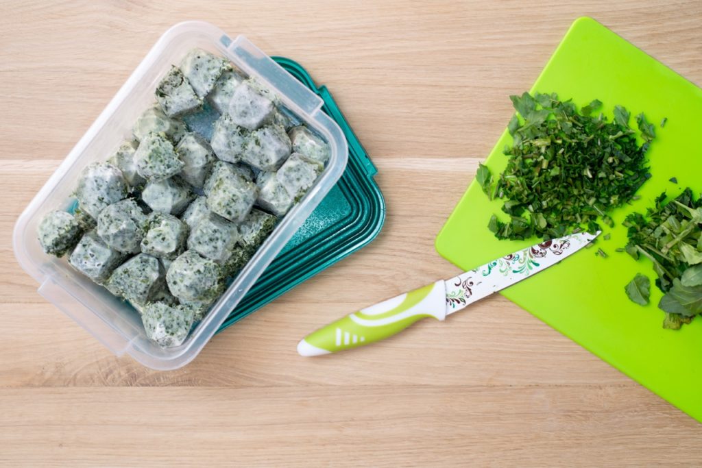 frozen ice cubes full of herbs in a Tupperware box next to a chopping board with chopped up herbs on its surface