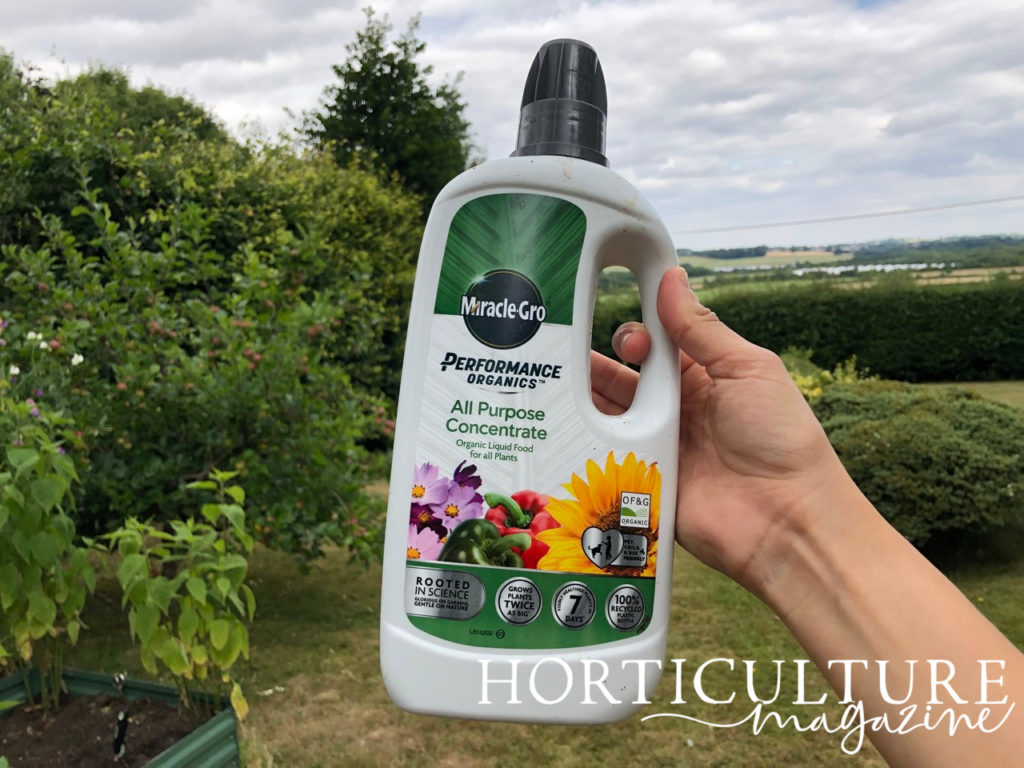 someone holding up a bottle of organic fertiliser in front of a garden full of hedges and trees on a cloudy day