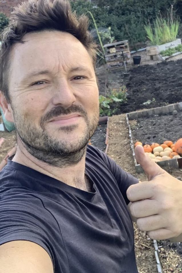 Joe Harrison smiling and giving a thumbs up with his vegetable plots in the background