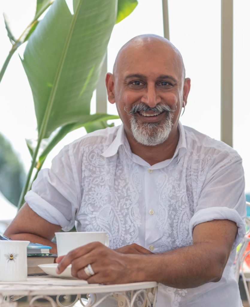 Manoj Maldé smiling as he sits at a table indoors in front of a houseplant