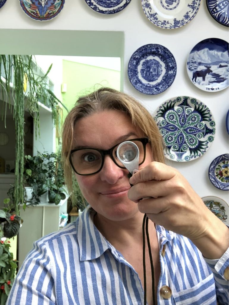 Jane Perrone holding up a magnifying glass to her eye with lots of plates and houseplants in the background