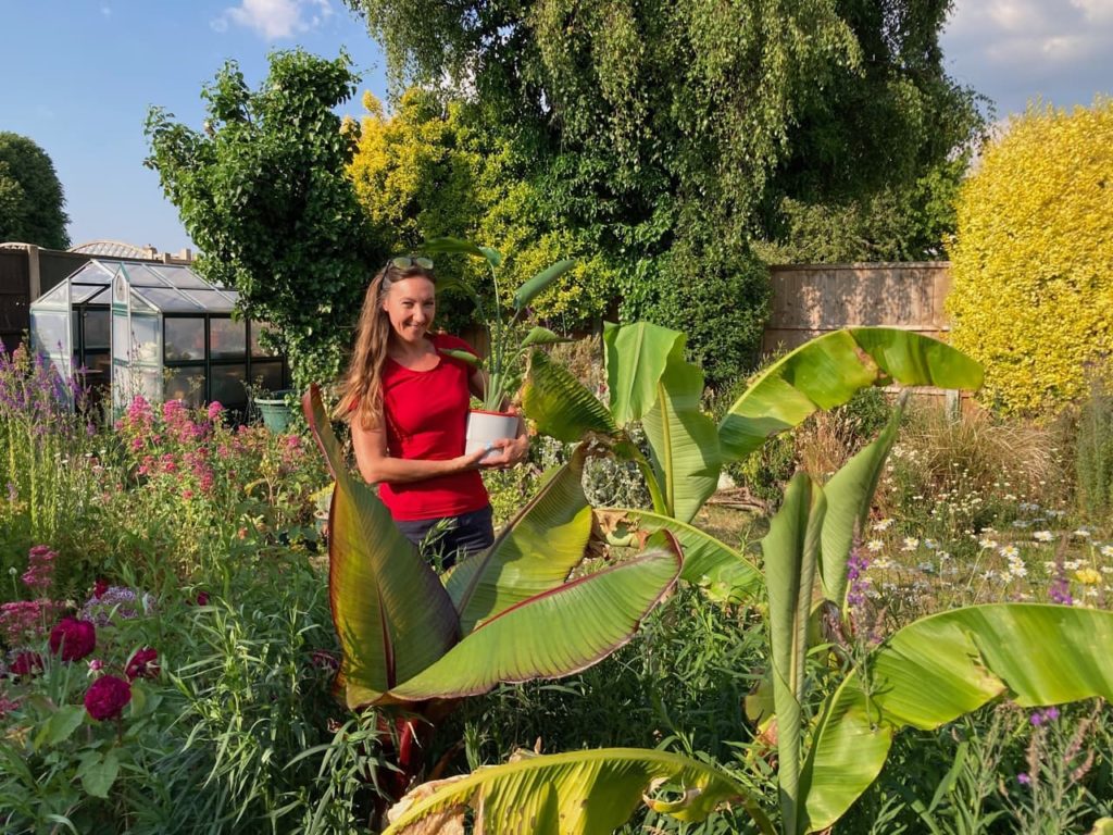 Debi holding up a potted plant in a garden surrounded by various colourful and tropical plants