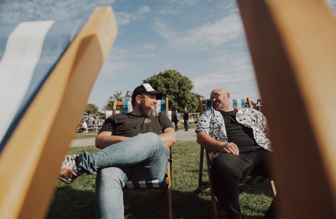 Chris and JJ sitting on deck chairs chatting outside at a festival