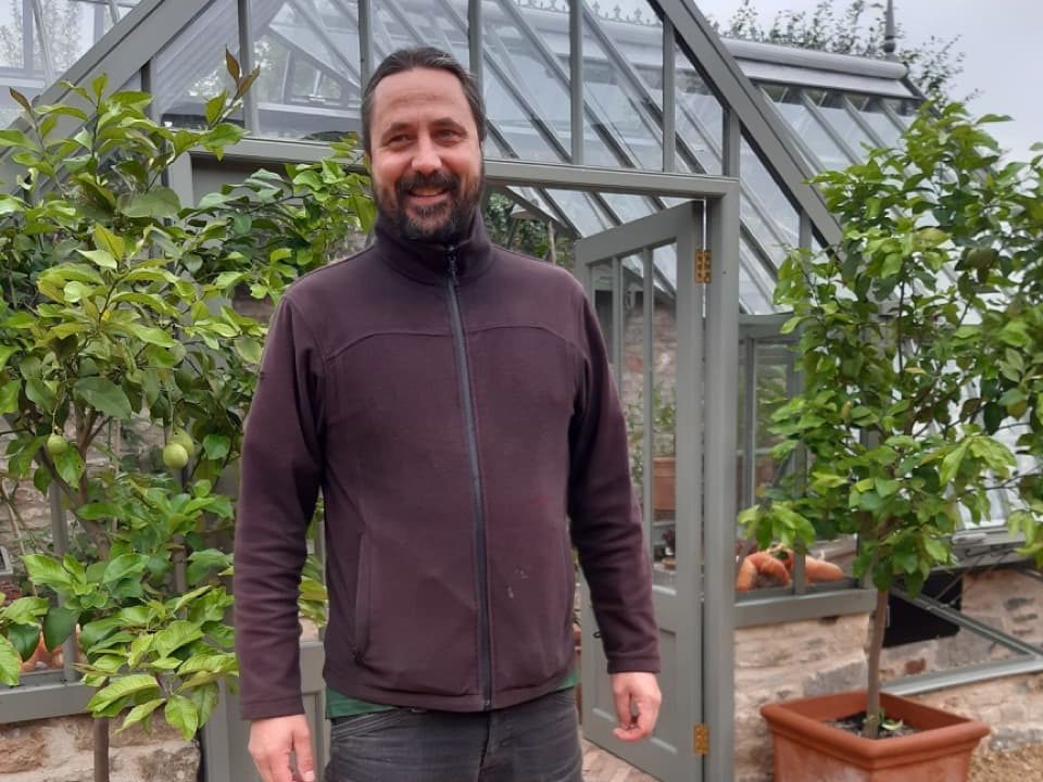 a greenhouse and potted pear trees in the background of a photo of Elliott Beveridge