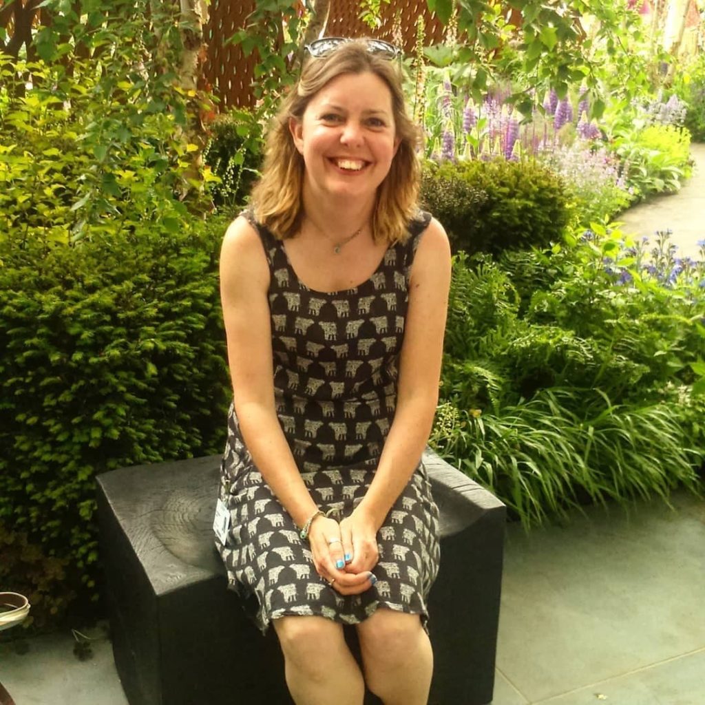 Nic Wilson sat on a black block outside in a beautiful garden, with various plants in the background