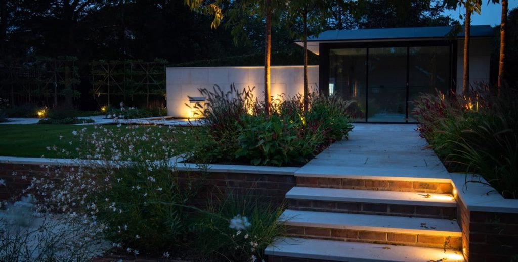 a modern garden at night full of beds and borders with lights along the path and on the lawn