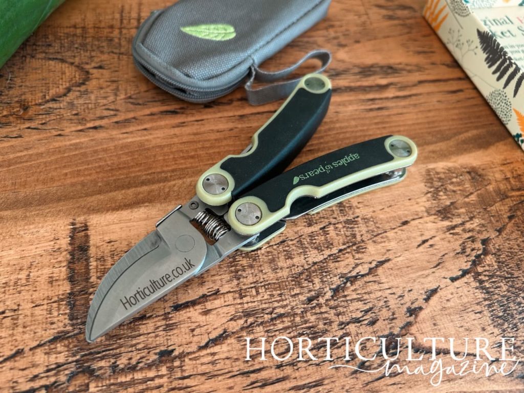 personalised secateurs lying on a wooden surface next to a grey pouch for storing them with 'horticulture.co.uk' engraved onto the blade