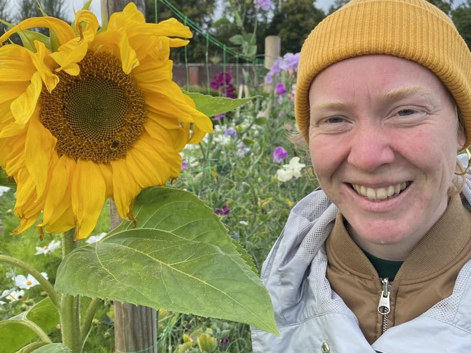 Gemma Jerome in her allotment with a large sunflower that is as tall as she is