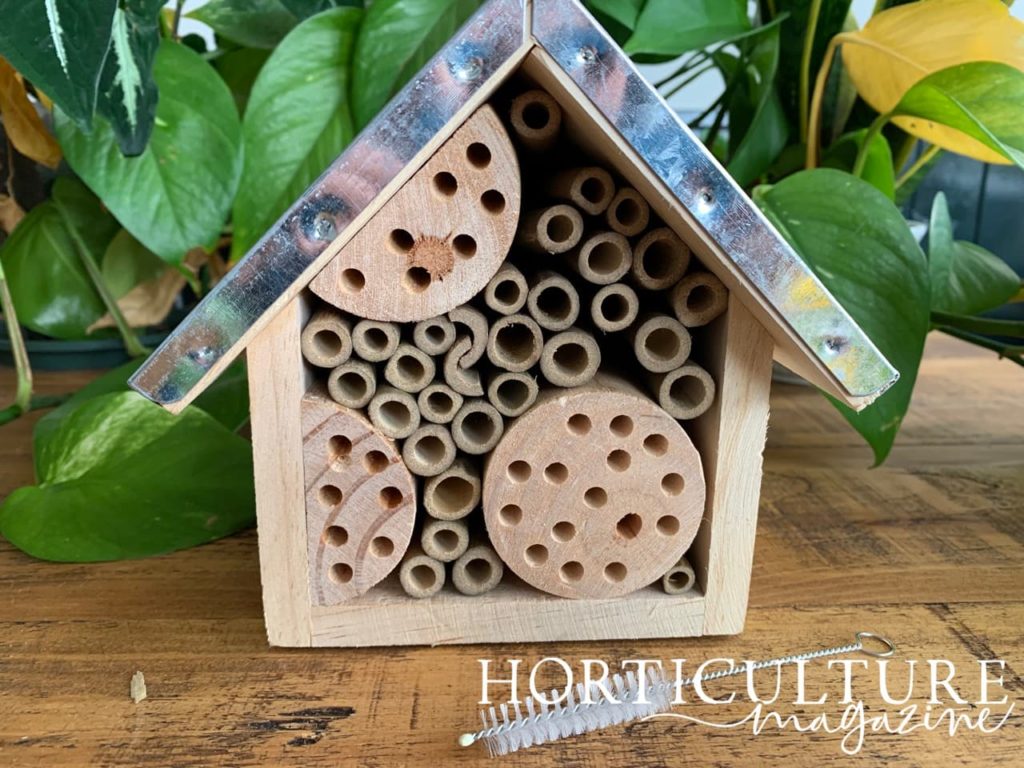 metal roof on a pentagonal-shaped bug hotel with wooden blocks and bamboo holes, with accompanying cleaning brush