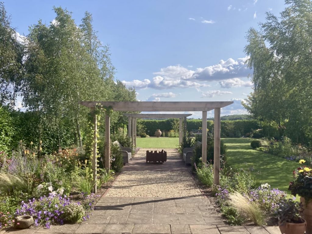 a stone pathway with a wooden pergola over the top surrounded by a lawn and flowering edges